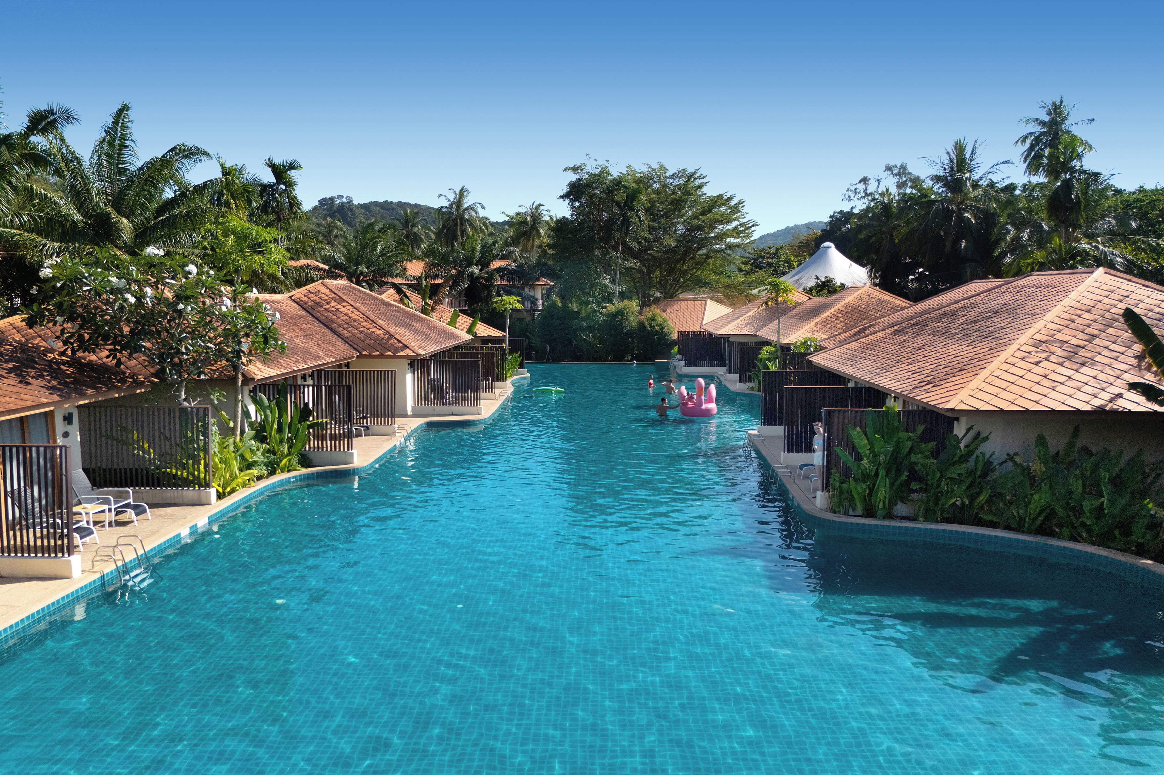 A swimming pool with a clear blue water, surrounded by palm trees and buildings with tiled roofs.