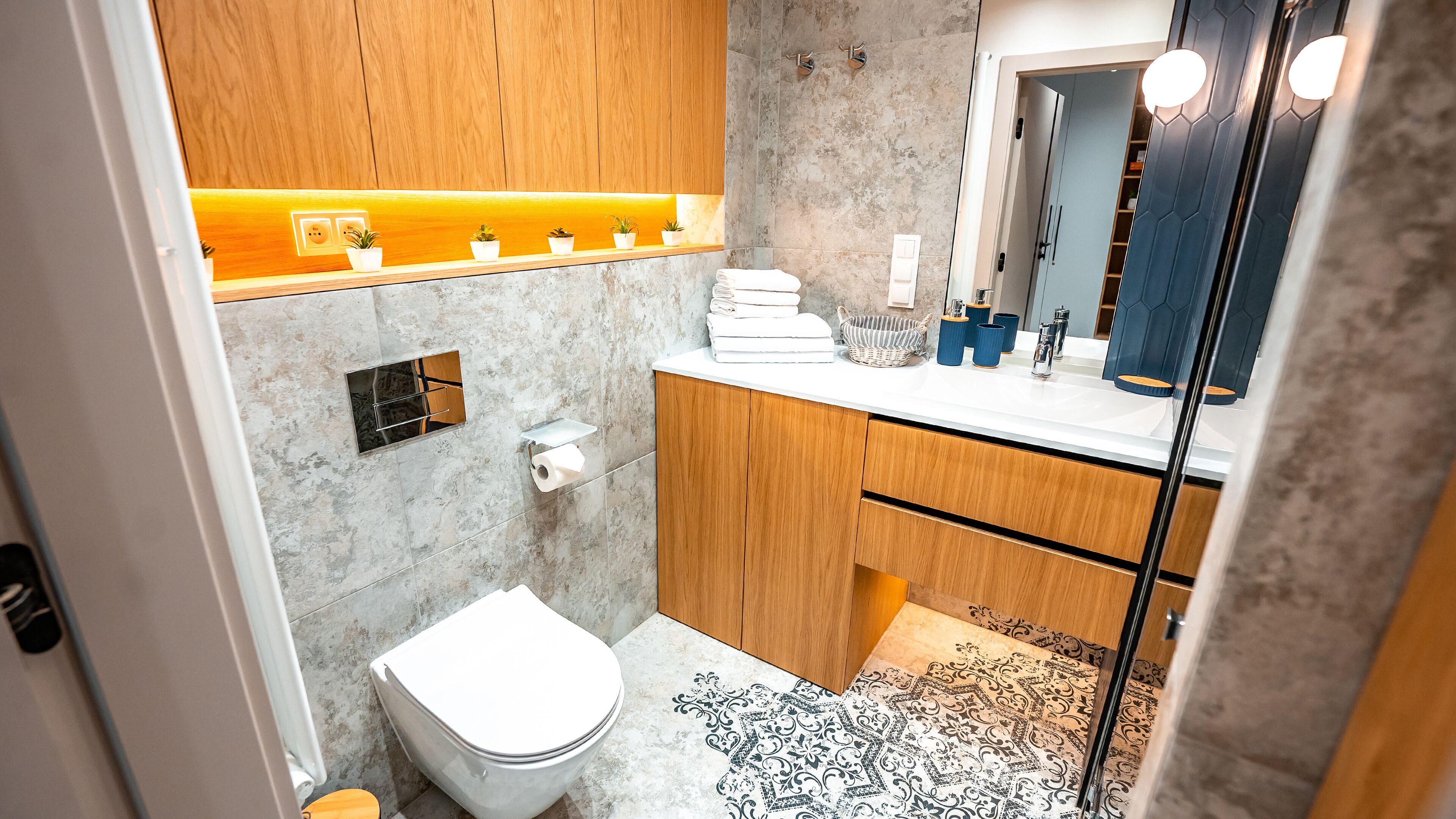 A modern bathroom with a marble wall, wooden vanity, and a large mirror.