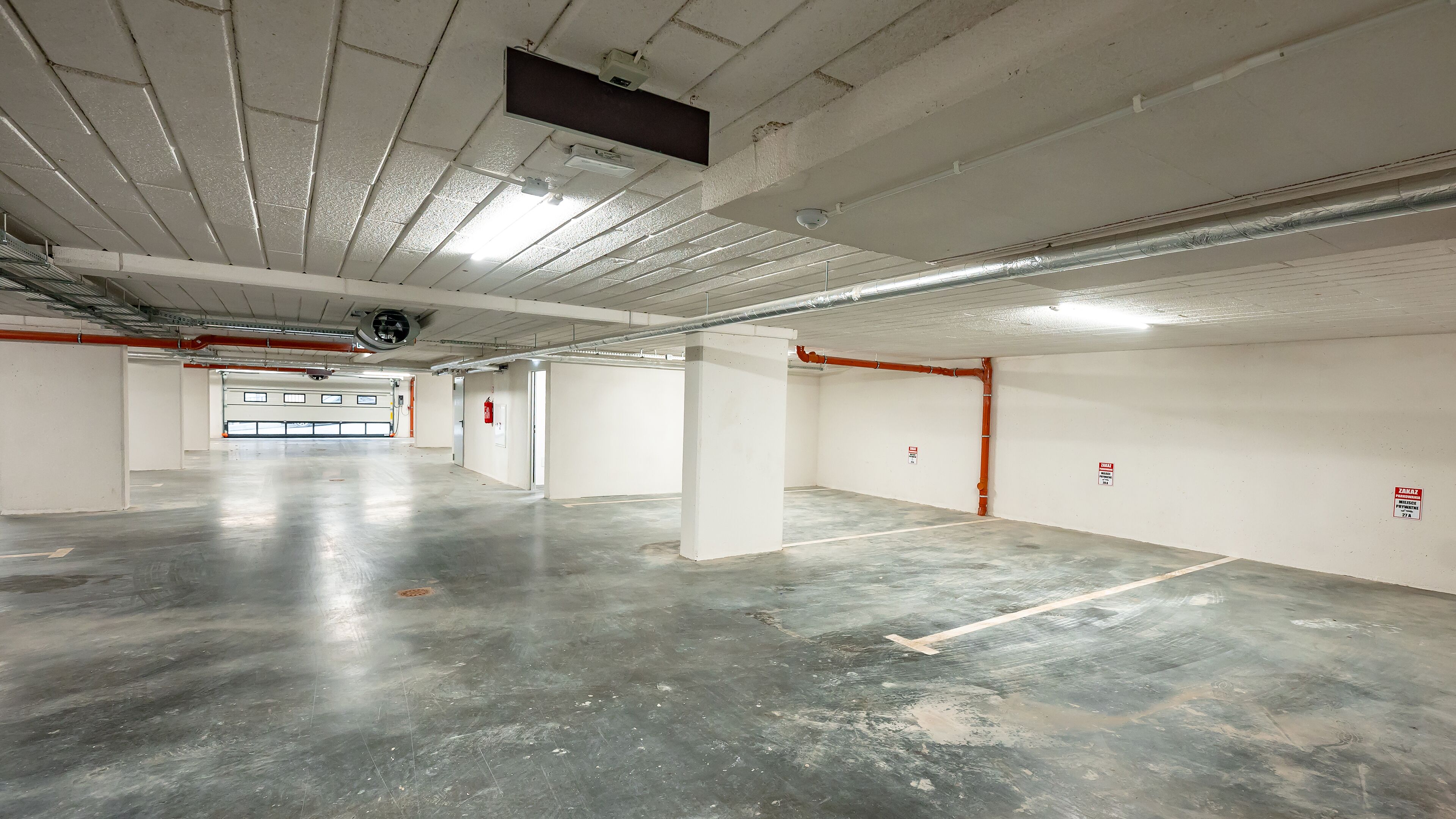 A large, empty parking garage with concrete floors and white walls, featuring several pillars, emergency exit signs, and visible ductwork.