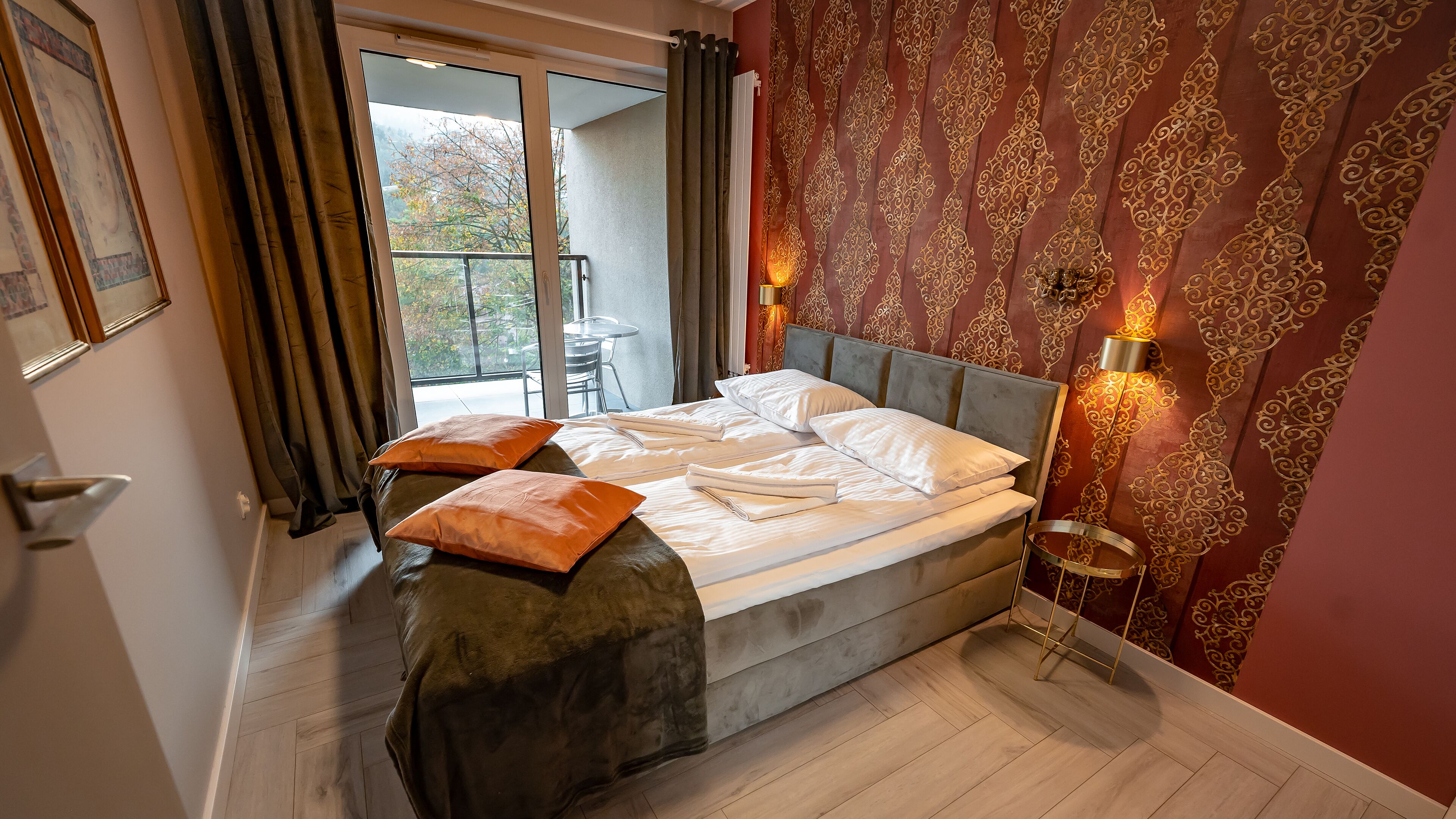 Un lit bien fait, avec des oreillers, un plancher en bois, un balcon avec une chaise et un papier peint à motifs.