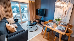 A modern living room with a sofa, coffee table, and dining table set for four. A TV is mounted on the wall, and there is a view of a snowy landscape through the windows. - Apartamenty Sun & Snow Cicha (Krynica-Zdroj)