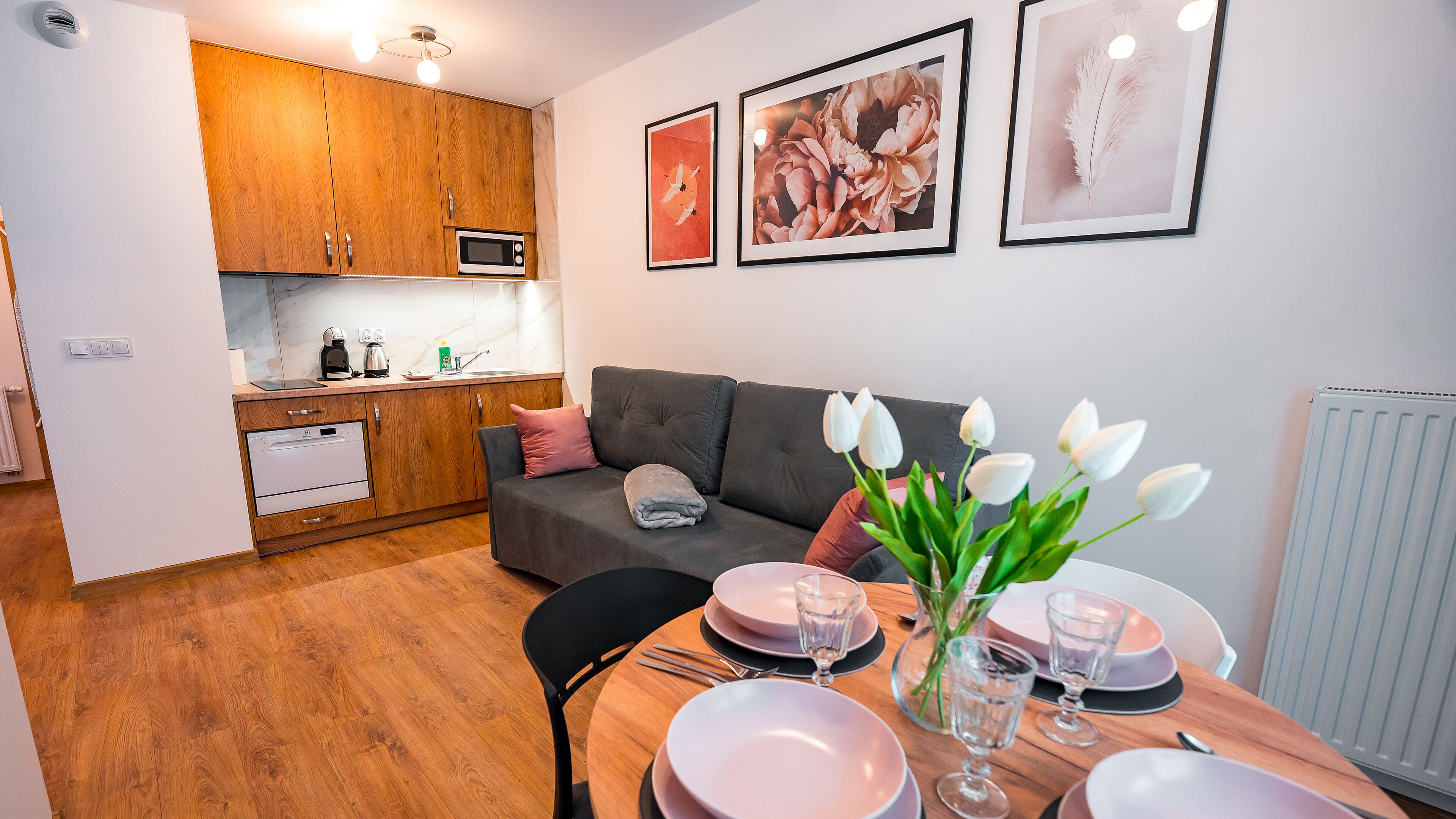 Une cuisine moderne avec des meubles en bois, un four à micro-ondes et un évier. On y trouve également un coin repas avec une table dressée pour deux personnes, un canapé et un vase rempli de fleurs blanches.
