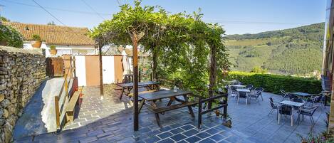 Un patio con suelo de piedra, mesas de madera y una pérgola cubierta de vegetación.