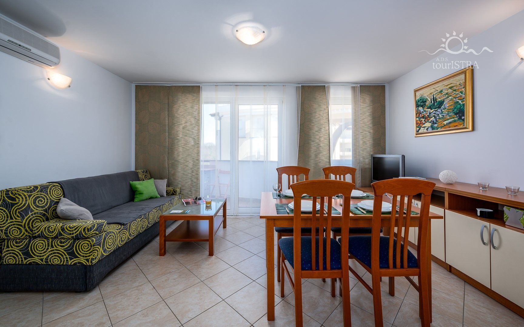 A modern living room with a sofa, dining table, and chairs. There is a TV and a painting on the wall.