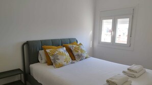 A neatly made bed with white linens and two pillows, a small bedside table, and a window with blinds. - Feel Welcome Barcelona Splau (Cornella de Llobregat)