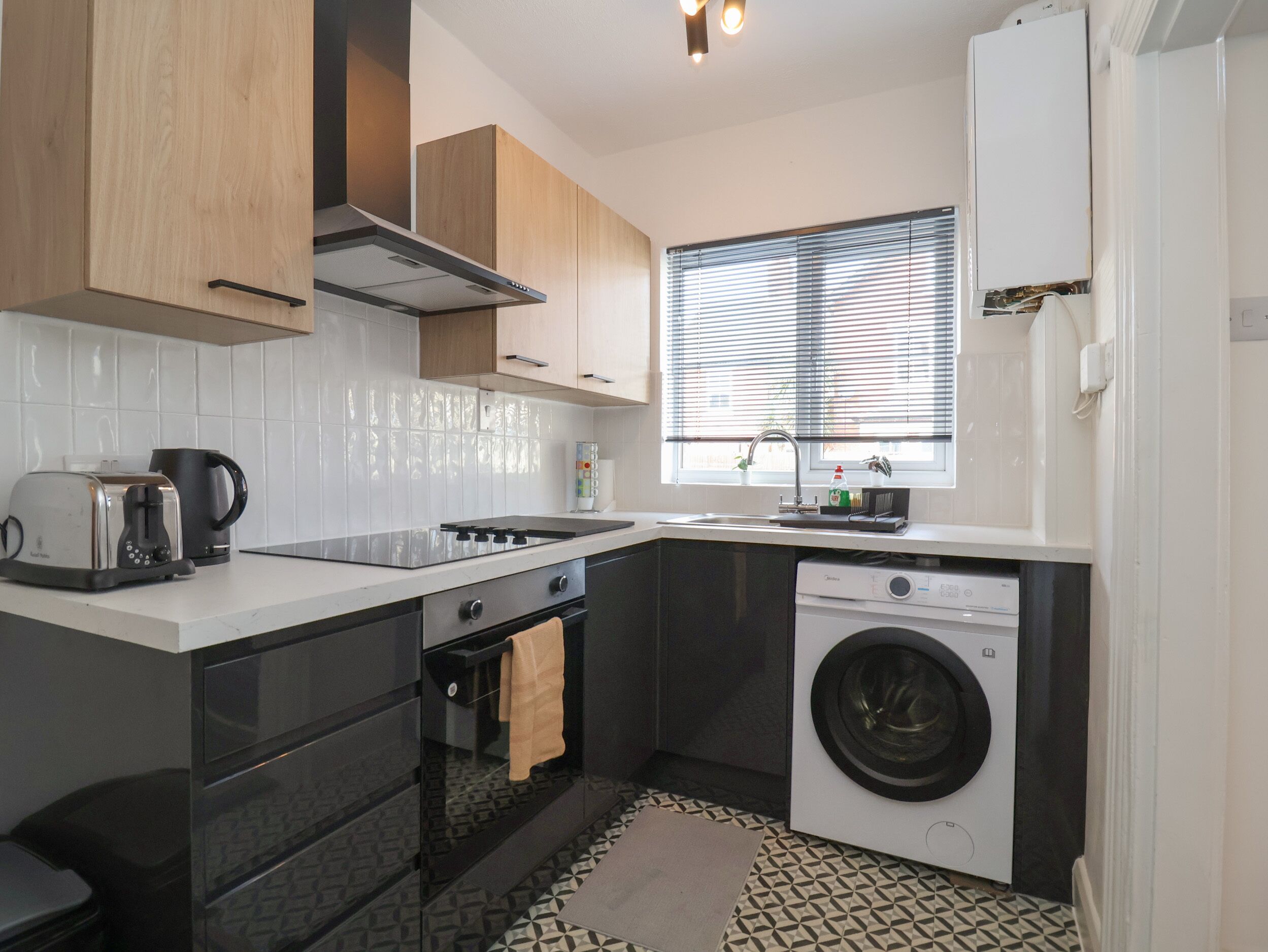 A compact kitchen with a built-in oven, a washing machine, and a window with blinds.