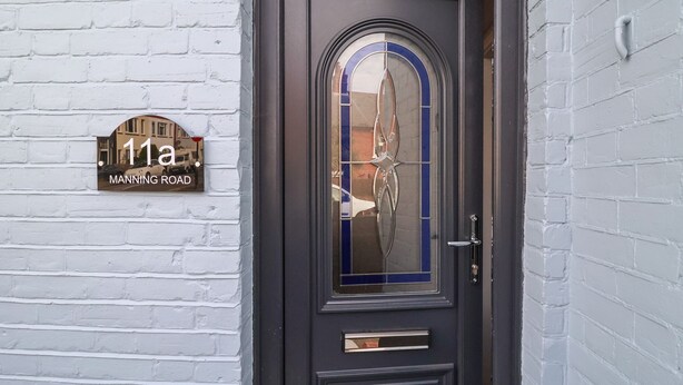 A dark-colored door with a glass panel and a brass handle, a white brick wall, and a house number sign indicating "11a Manning Road".