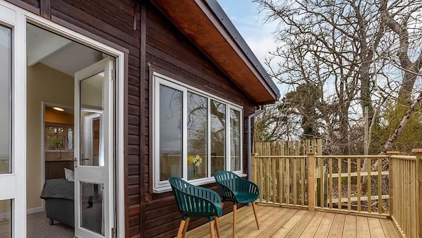 A wooden deck with two green chairs, a wooden fence, and a building with large windows. - Kilninver Chalet on Reelig Estate (Inverness)