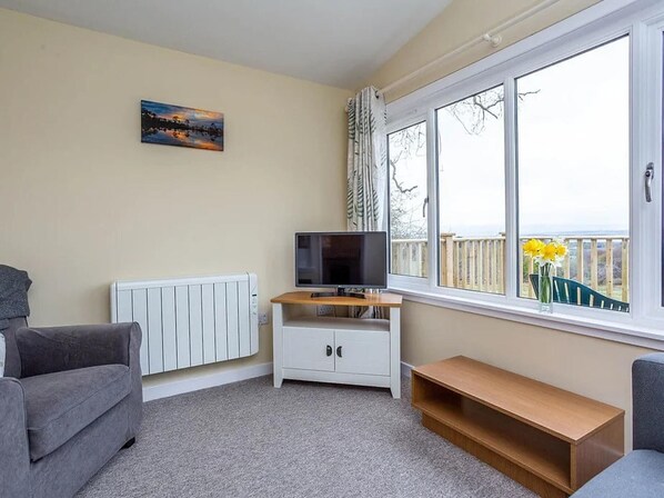 A living room with a white radiator, a TV on a wooden stand, a grey sofa, a wooden bench, and a window with a view of a deck and yellow flowers.