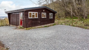 A small, single-story wooden cabin with a gravel driveway, surrounded by trees and a wooden fence. - Kilninver Chalet on Reelig Estate (Inverness)