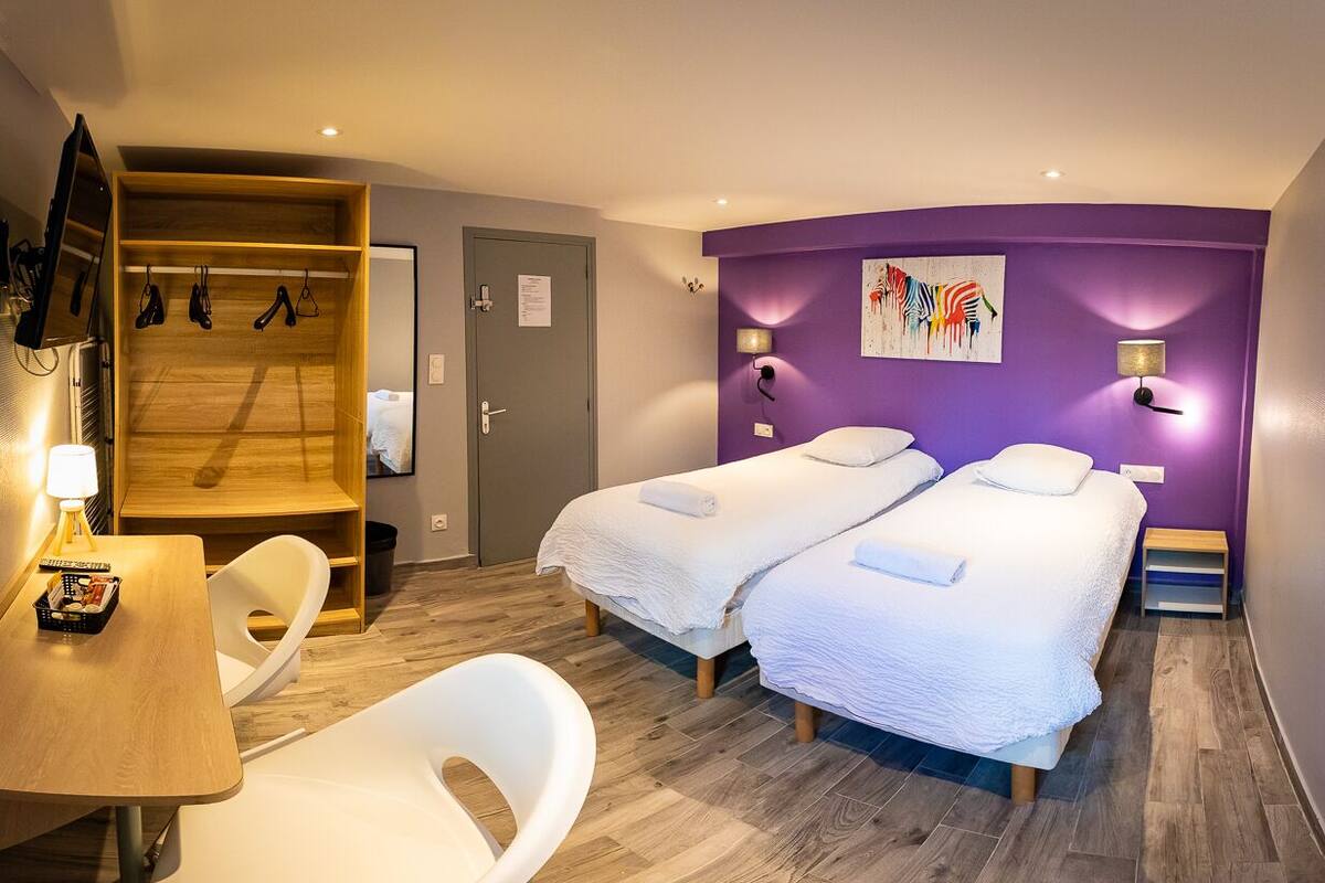 A hotel room with two beds, a wooden wardrobe, a desk with a chair, and a small table with a lamp.