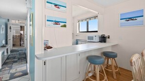 Een moderne keuken met witte kasten, een wit aanrecht en twee blauwe barstoelen. Op de muur hangen drie foto’s in lijsten met afbeeldingen van boten en waterlandschappen.