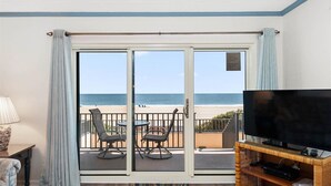Ein Zimmer mit einer großen Schiebeglastür, die auf einen Balkon mit Blick auf den Strand führt. Auf dem Balkon befinden sich zwei Stühle und ein Tisch, im Zimmer ein Flachbildfernseher auf einem Holzboden.