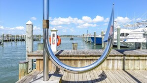 Un muelle de madera con una estructura metálica, un flotador salvavidas y barcos al fondo.
