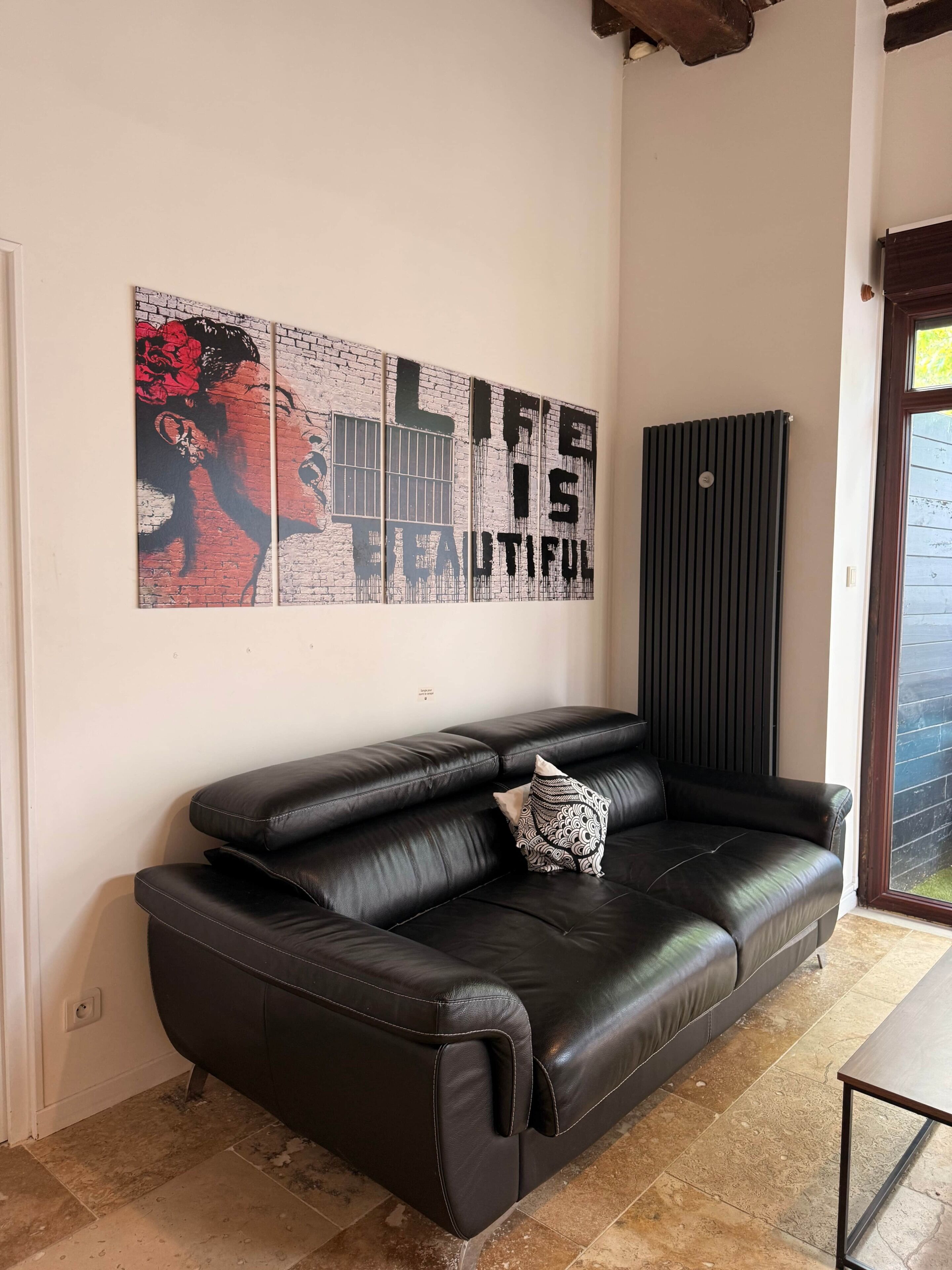 Un divano in pelle nera con un cuscino decorativo, una parete con un quadro raffigurante 'Life is Beautiful' e un termosifone.