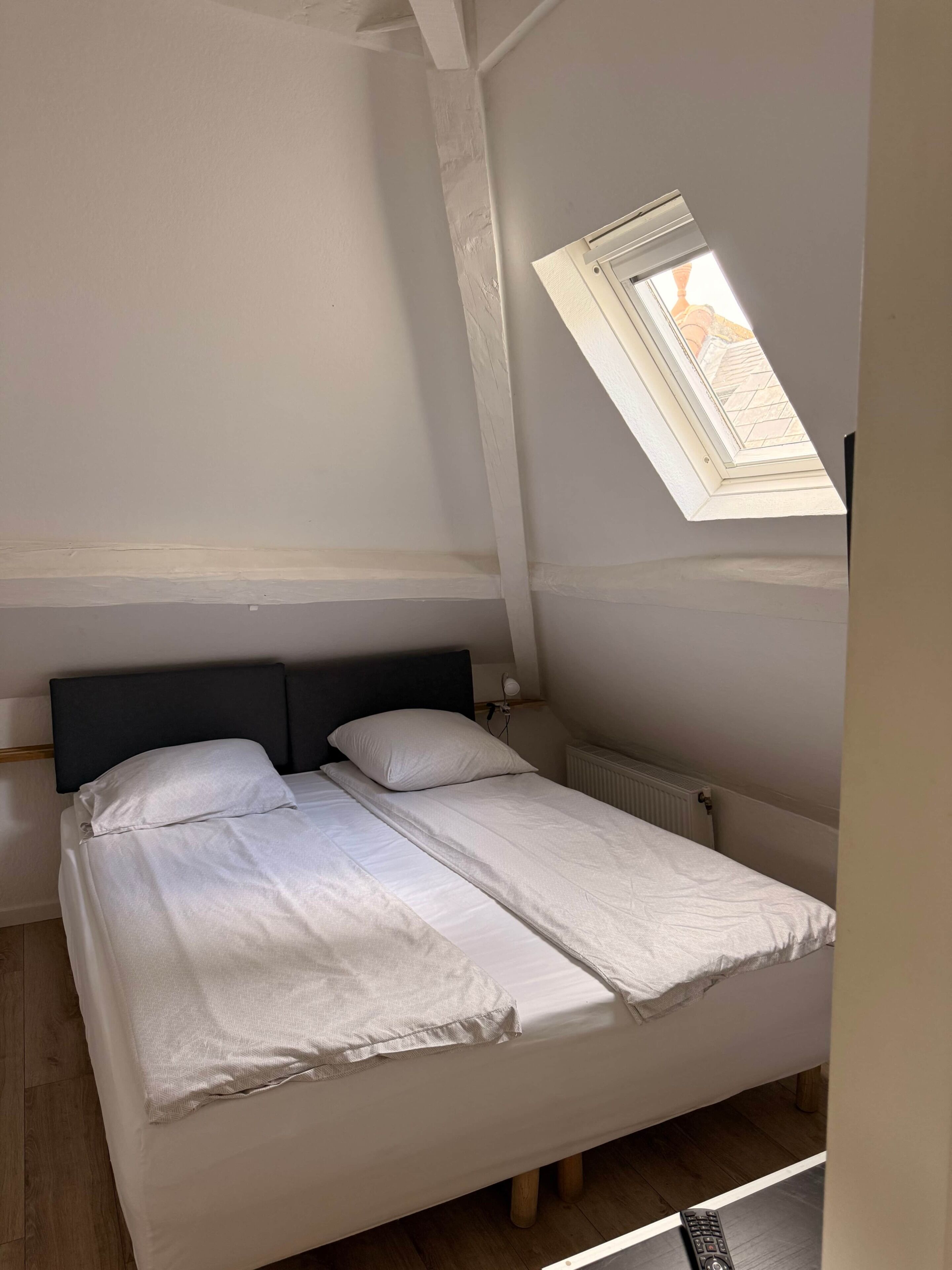 A single bed with a dark headboard, a white mattress, and a light-colored blanket. There is a window with a view of a building outside.