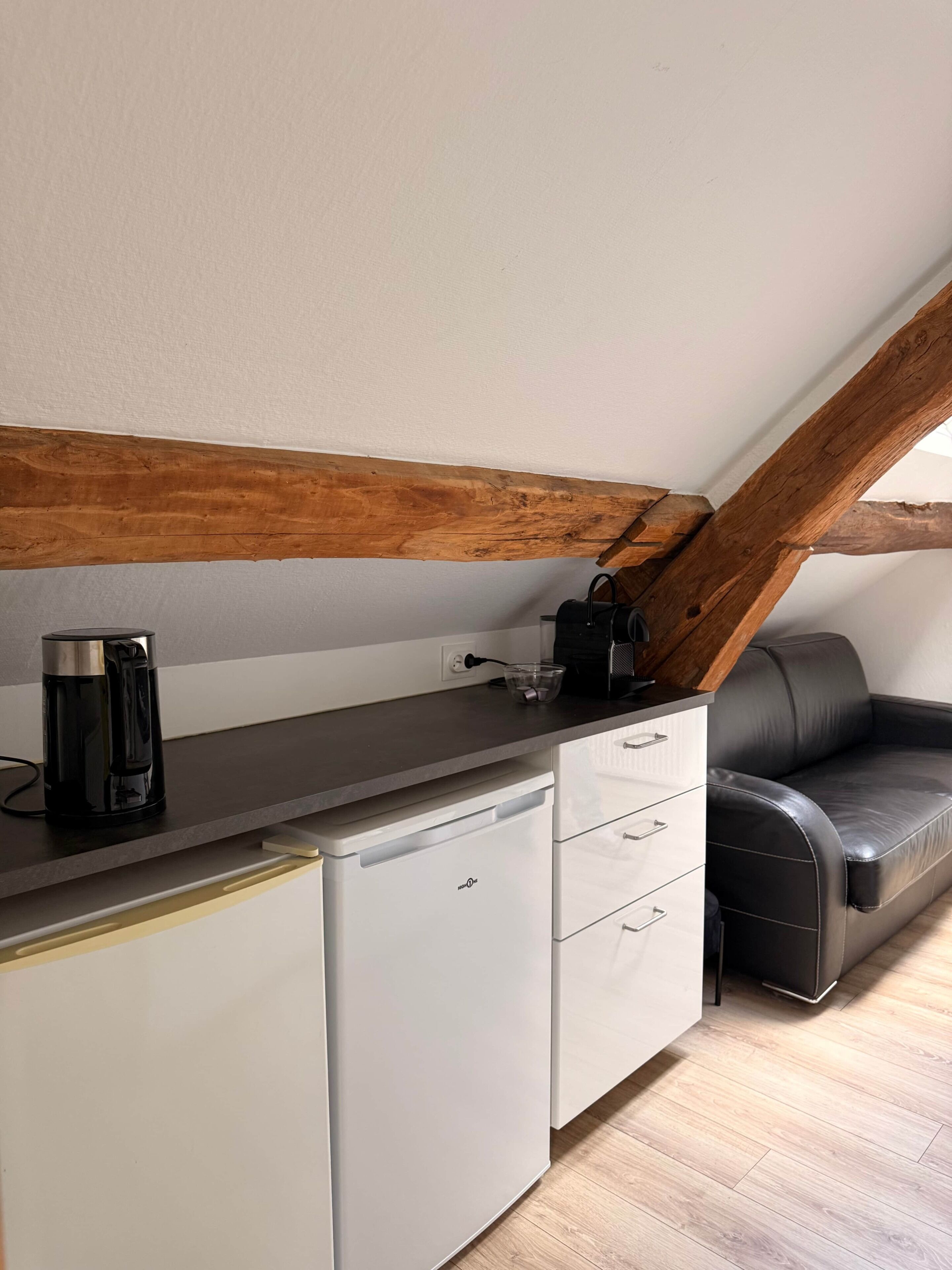 Una kitchenette con cafetera, hervidor y tostadora. Hay un refrigerador blanco, una encimera negra y un sofá de cuero negro. La habitación tiene vigas de madera en el techo.