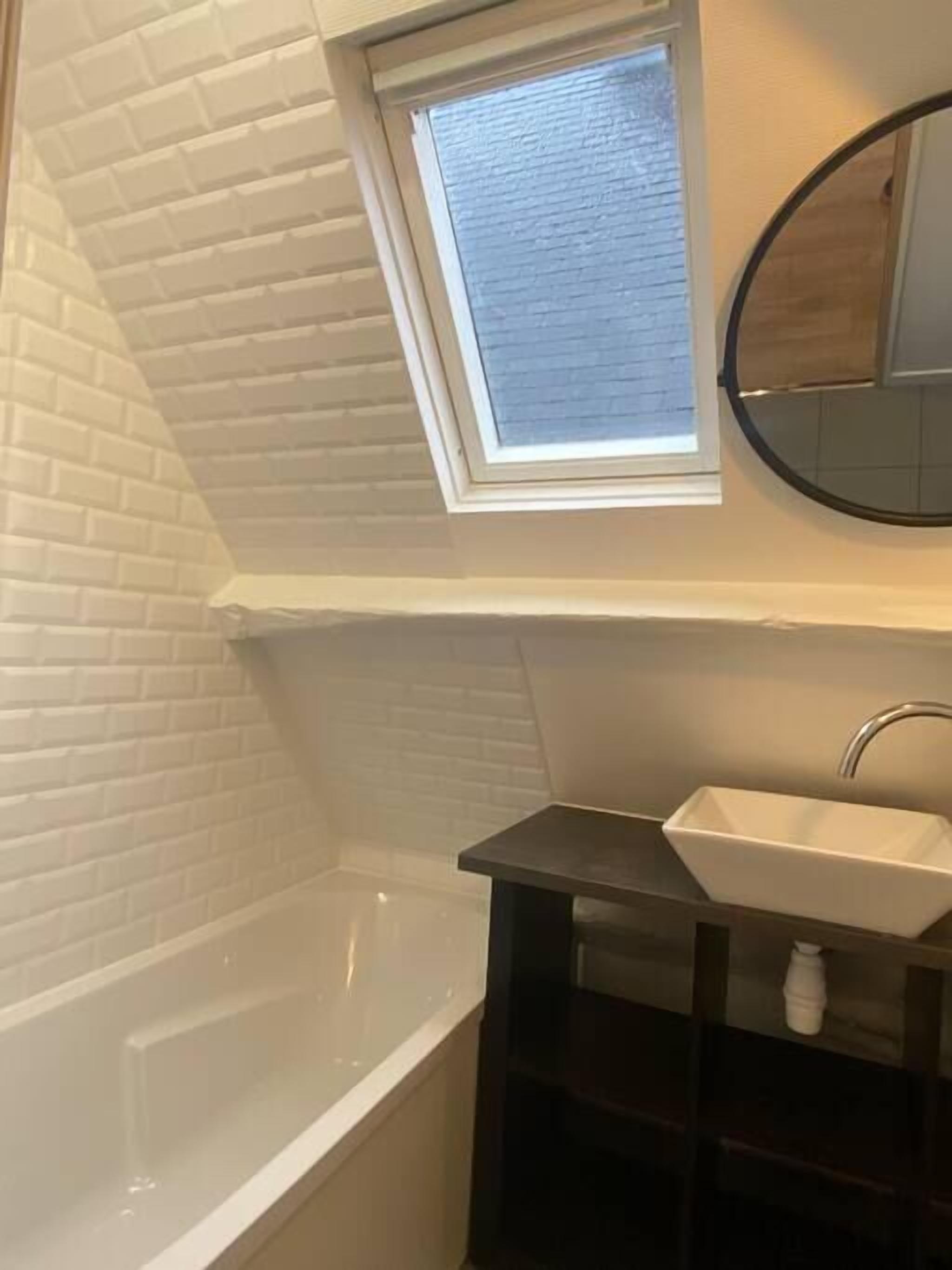 A bathroom with a white bathtub, a window above the sink, and a round mirror.