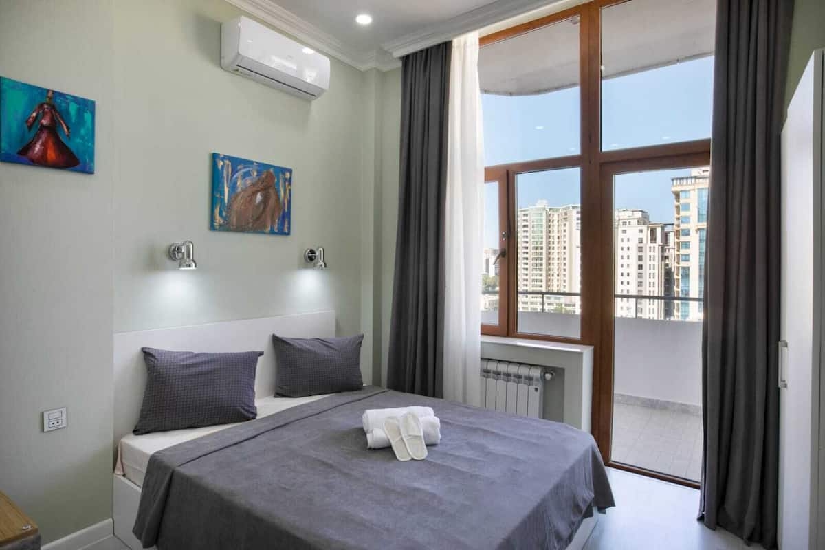 Ein Hotelzimmer mit einem Bett, zwei Kissen, einem Gemälde, einer Klimaanlage, einer Heizung und Blick auf Gebäude durch ein großes Fenster.