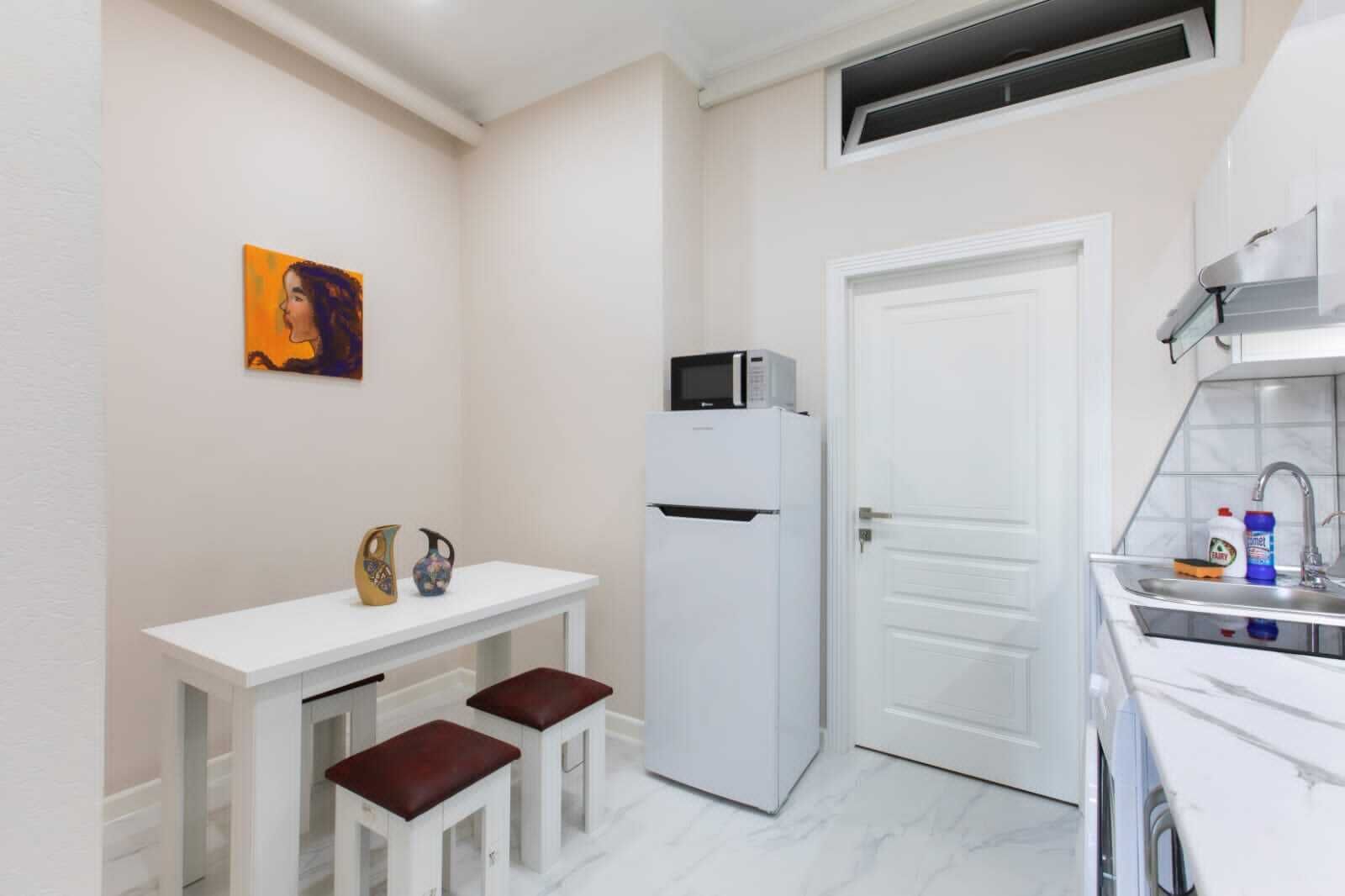 Une cuisine compacte comprenant un réfrigérateur blanc, un four à micro-ondes, un évier avec des produits de nettoyage et une petite table accompagnée de deux tabourets.