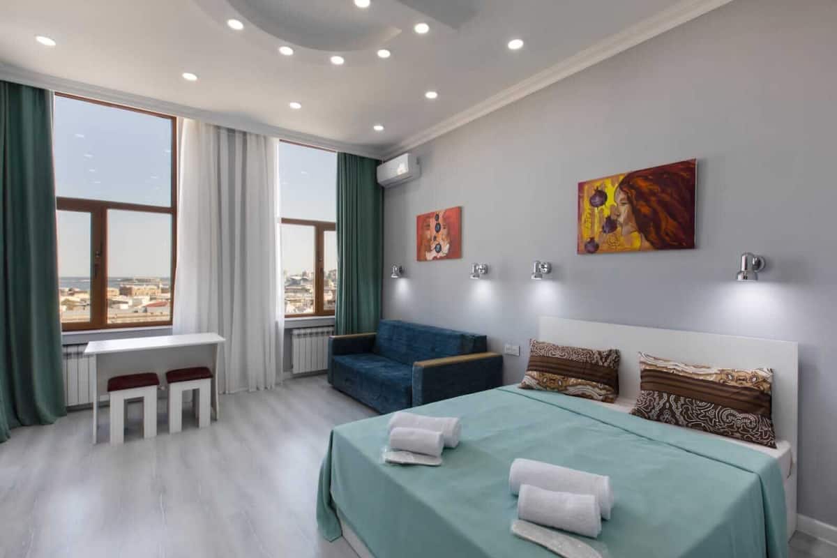 Ein modernes Hotelzimmer mit einem Bett, einem Schreibtisch und einer Couch. Große Fenster mit Vorhängen, Kunstwerke an den Wänden und Blick auf die Stadt.