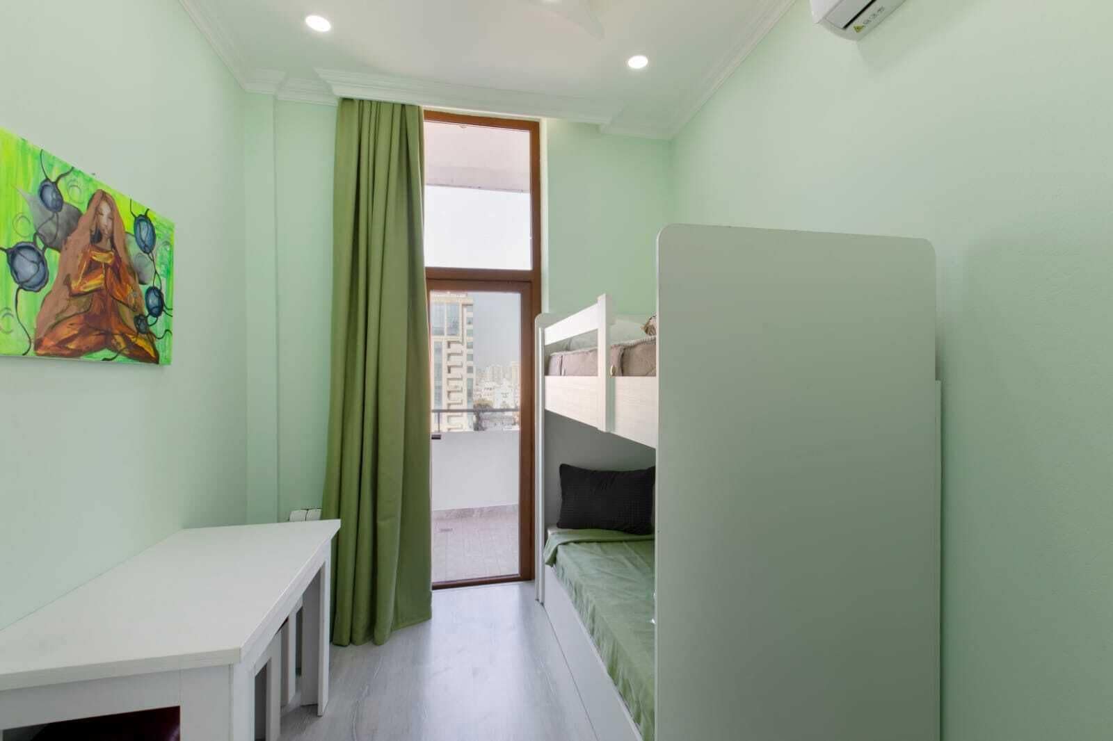 A small, well-lit room with a bed, a desk, and a window with curtains.