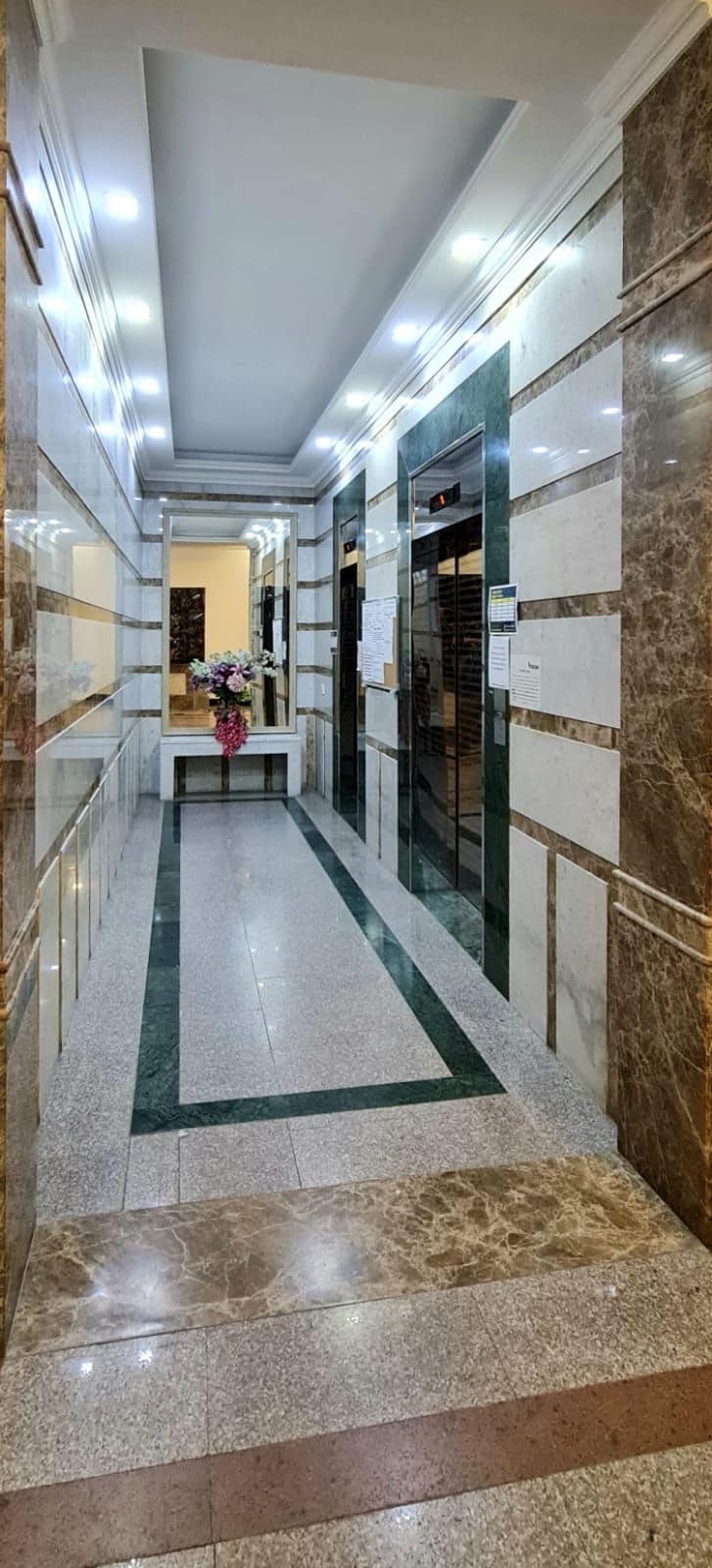 Un pasillo con piso de mármol, un arreglo floral central y un muro espejado.