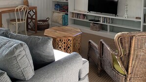 A cozy living room with a wooden coffee table, striped sofa, wicker chairs, a bookshelf, a television, and a chandelier. - Charming artist’s cottage near the beach, large lot with beautiful oaks (Oak Island)