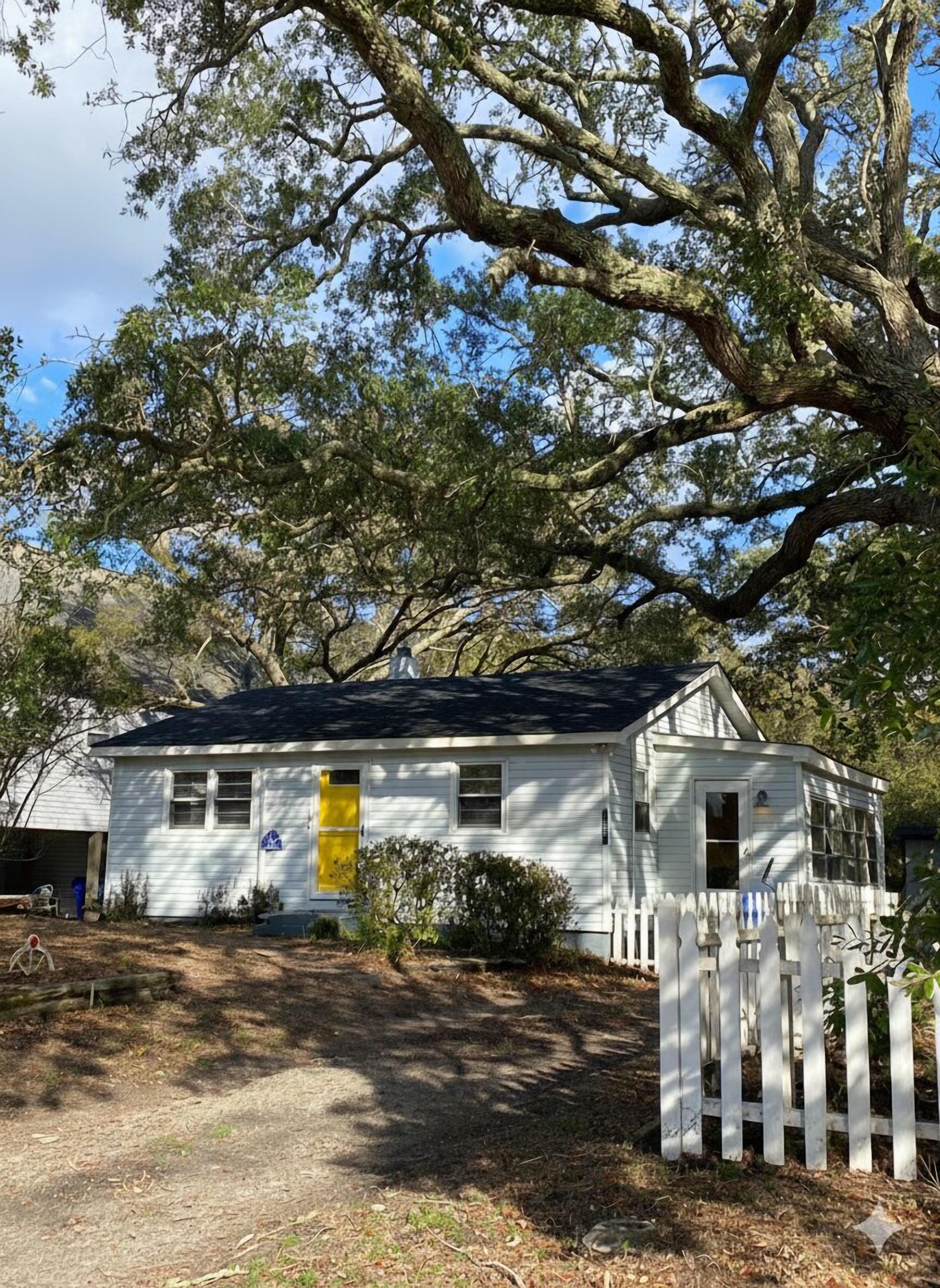 A white single-story house with a yellow door, surrounded by trees and a white picket fence.