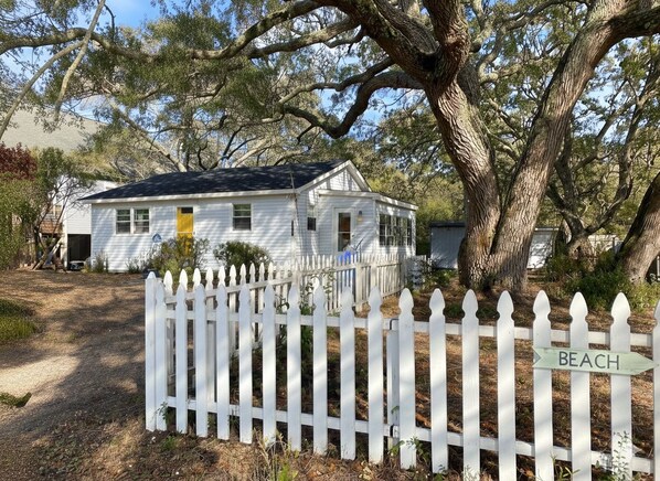 A white house with a porch, surrounded by a white picket fence, and a sign indicating 'BEACH' in the background.