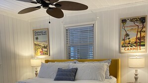 A bedroom with a bed, a ceiling fan, a window with blinds, and two wall-mounted lamps. - Charming artist’s cottage near the beach, large lot with beautiful oaks (Oak Island)