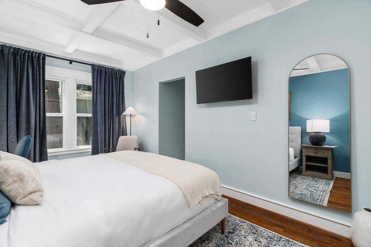 Un dormitorio con cama, ventilador de techo, televisor, espejo y ventana con cortinas.