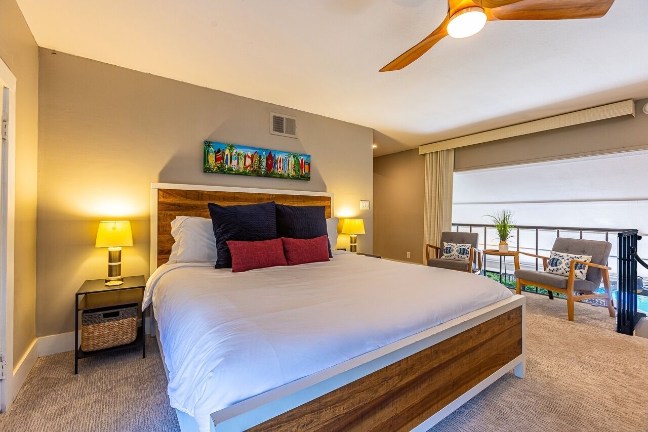 A hotel room with a large bed, two bedside tables with lamps, a ceiling fan, a balcony with chairs, and a colorful painting above the bed.