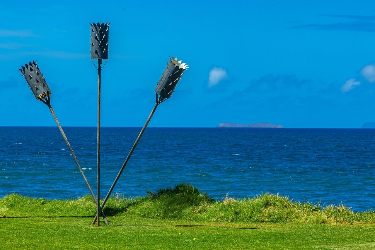 Une sculpture composée de trois objets hauts et pointus, installée dans un espace verdoyant, d’où l’on peut admirer l’océan et une île lointaine.