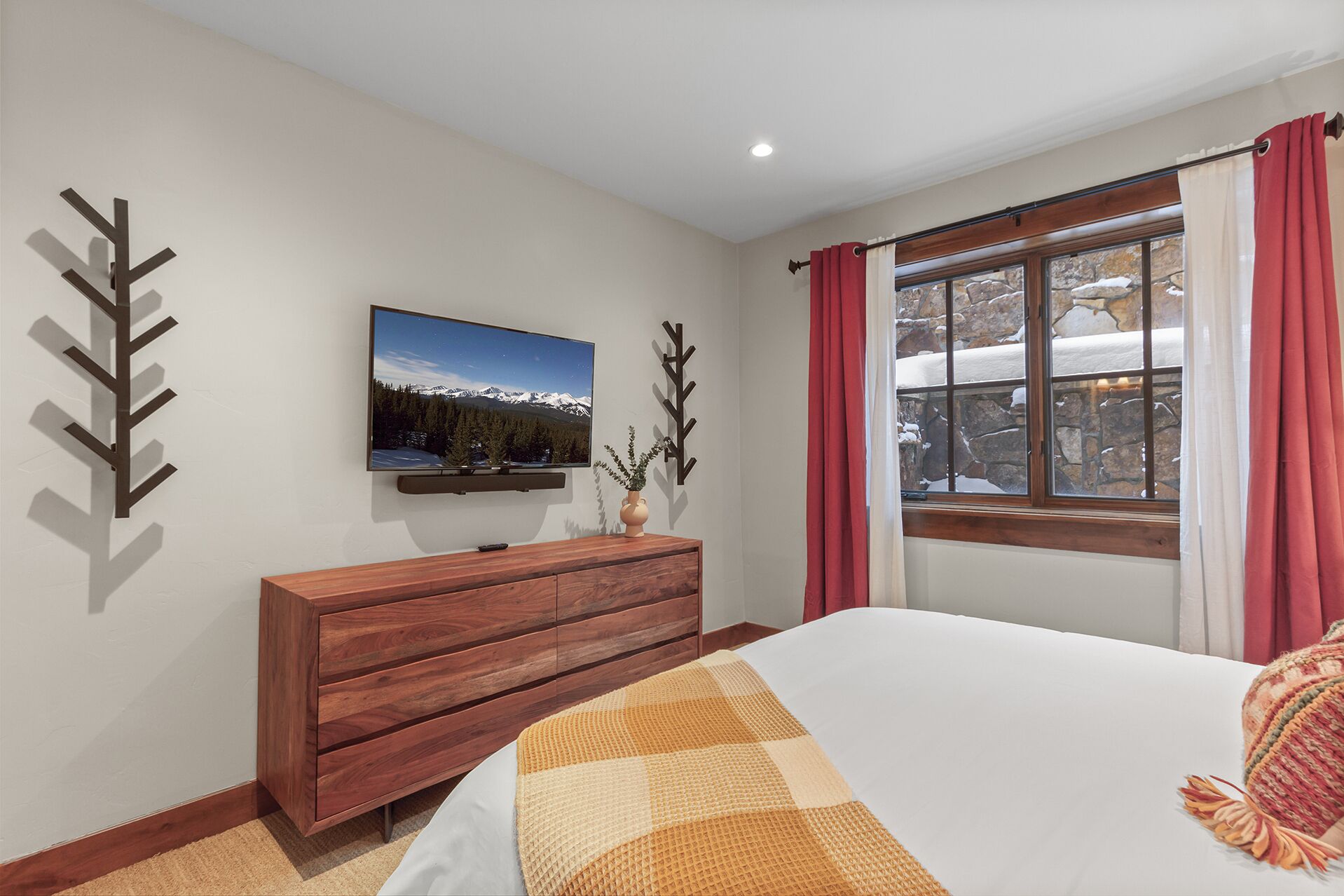 A bedroom with a bed, a wooden dresser, a flat-screen TV, a window with curtains, and a wall-mounted plant.