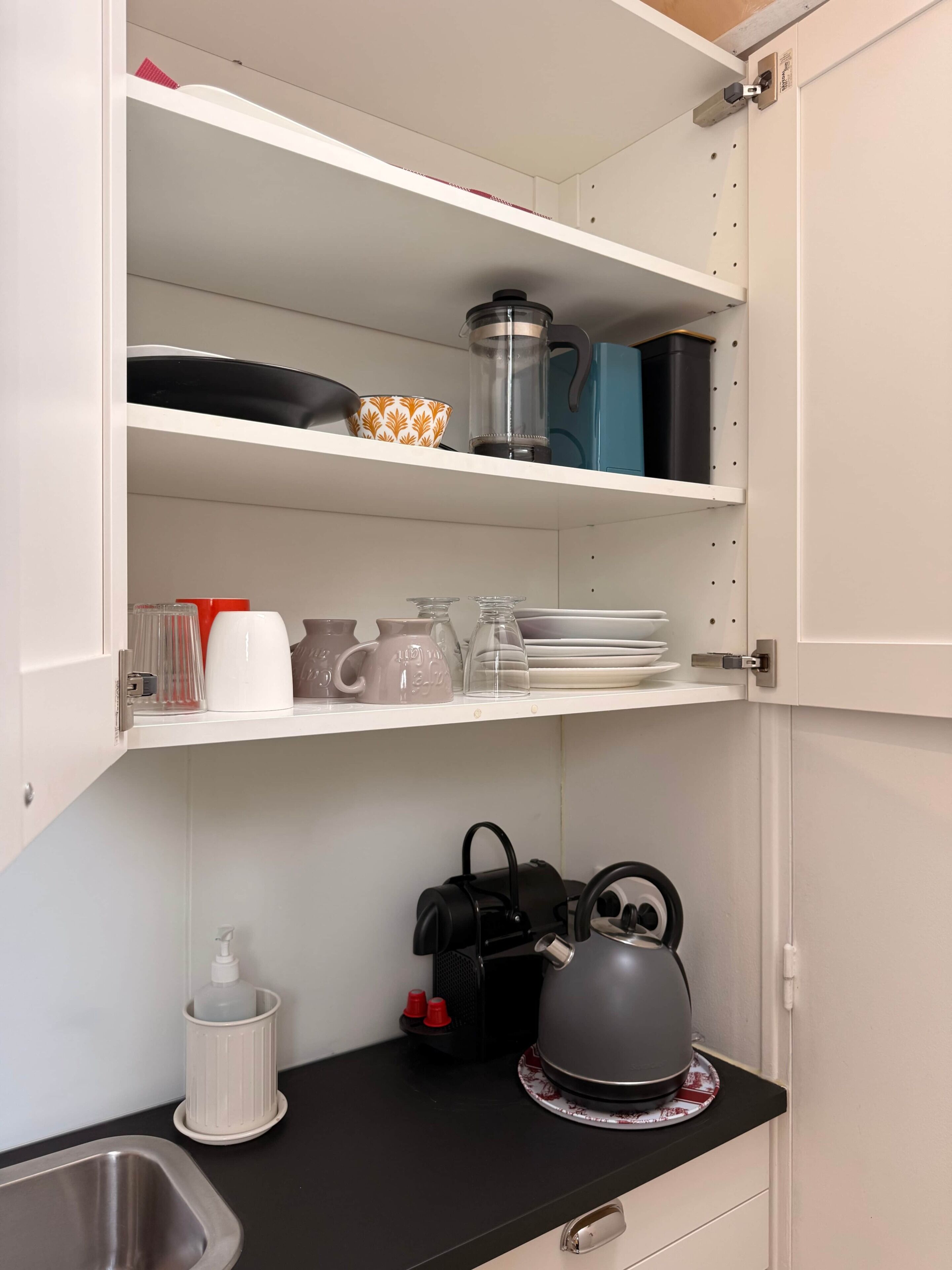 Een keuken met een zwart aanrecht, een waterkoker, een koffiezetapparaat en verschillende borden en glazen op de planken.