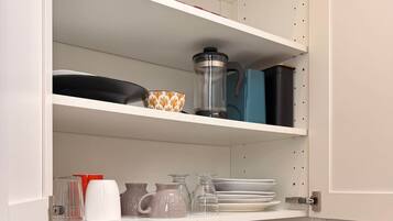 Une cuisine avec un plan de travail noir, une bouilloire, une machine à café, ainsi que divers plats et verres rangés sur les étagères.
