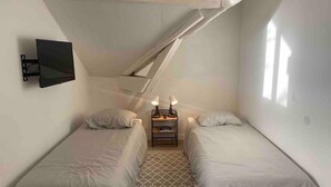 Une petite pièce bien éclairée, avec deux lits simples, une table de chevet avec une lampe et un téléviseur fixé au mur.