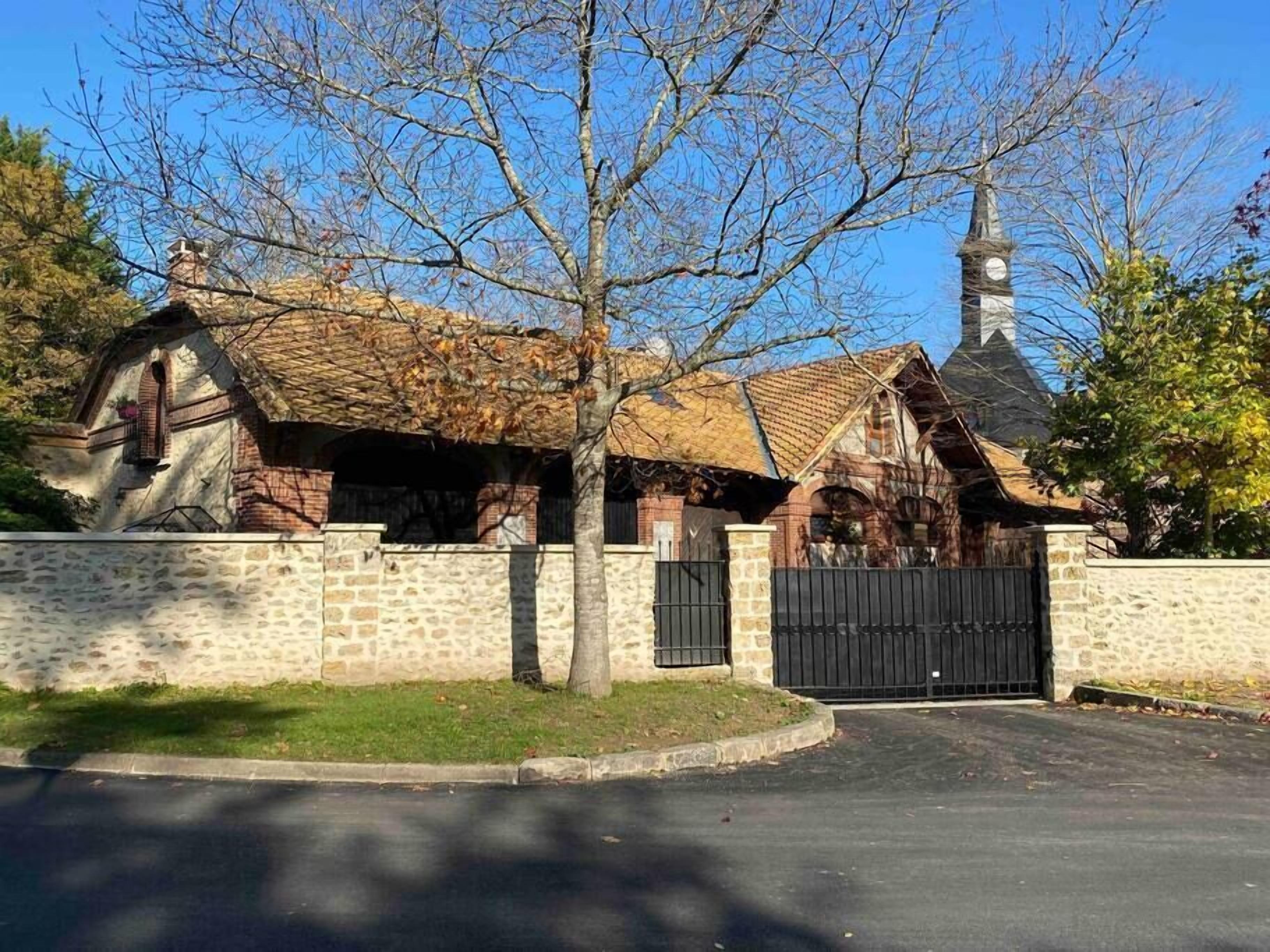 Un bâtiment en pierre avec un toit en briques, une porte noire et, au fond, un clocher.
