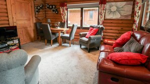 A cozy cabin interior with a wooden dining table, two grey chairs, a leather sofa with red cushions, and a television on a stand. - 10 Water's Edge (Morpeth)