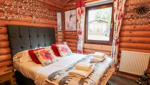 A log cabin bedroom with a large bed, floral pillows, a decorative wall piece, and a window with curtains. - 10 Water's Edge (Morpeth)