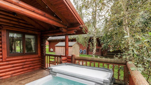 A wooden cabin with a hot tub on the deck, surrounded by trees. - 10 Water's Edge (Morpeth)