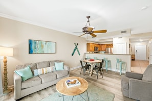 A living room with a sofa, coffee table, and a ceiling fan. There is a kitchen area with a bar and stools.