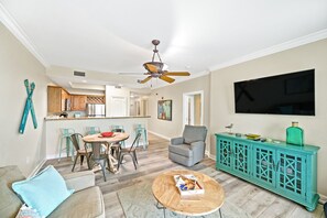 A living room with a sofa, a coffee table, a dining area with chairs, a kitchen in the background, and a teal cabinet with a TV mounted on the wall.