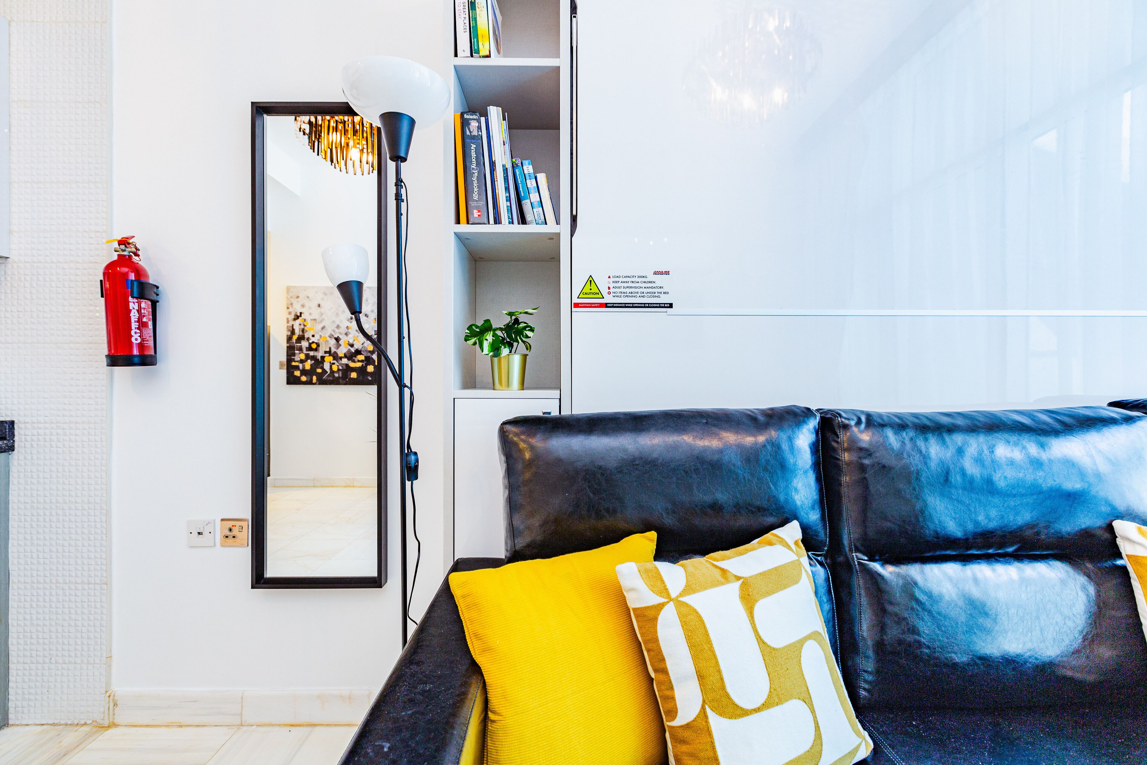 A modern living room with a black leather sofa, yellow and white cushions, a bookshelf, a mirror, and a fire extinguisher.