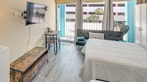 A hotel room with a bed, a desk, a chair, a TV mounted on the wall, and a balcony with curtains.
