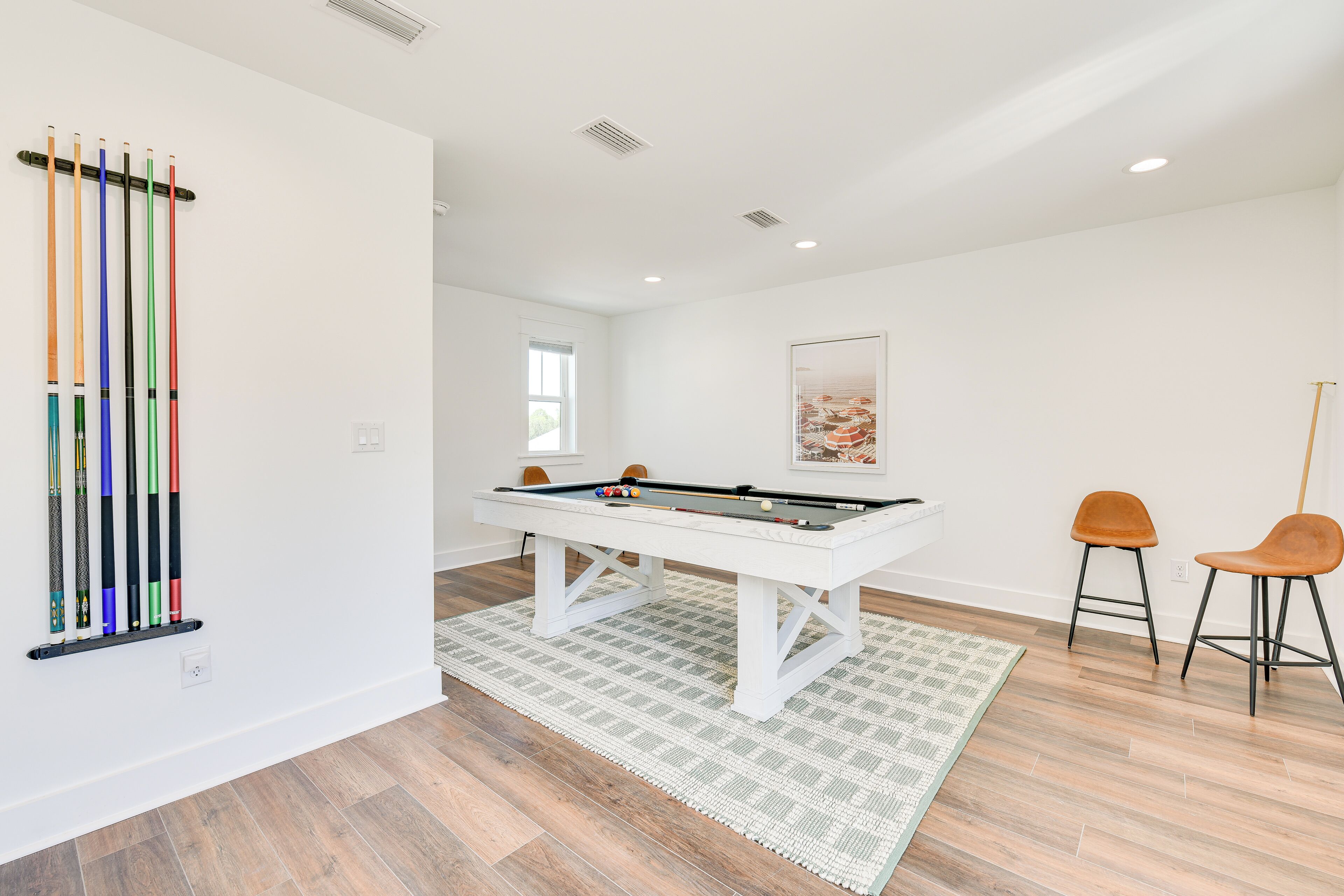 A pool table with a white frame, two orange barstools, and a pool cue rack against the wall.