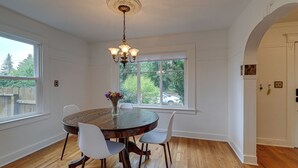 Uma sala de jantar com uma mesa redonda, cadeiras brancas, um candelabro e uma janela com vista para árvores e um carro.