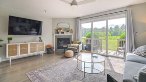 A modern living room with a flat-screen TV, a fireplace, a balcony with a view of greenery, and a ceiling fan. - North Conway Home for Fall Adventures (North Conway)