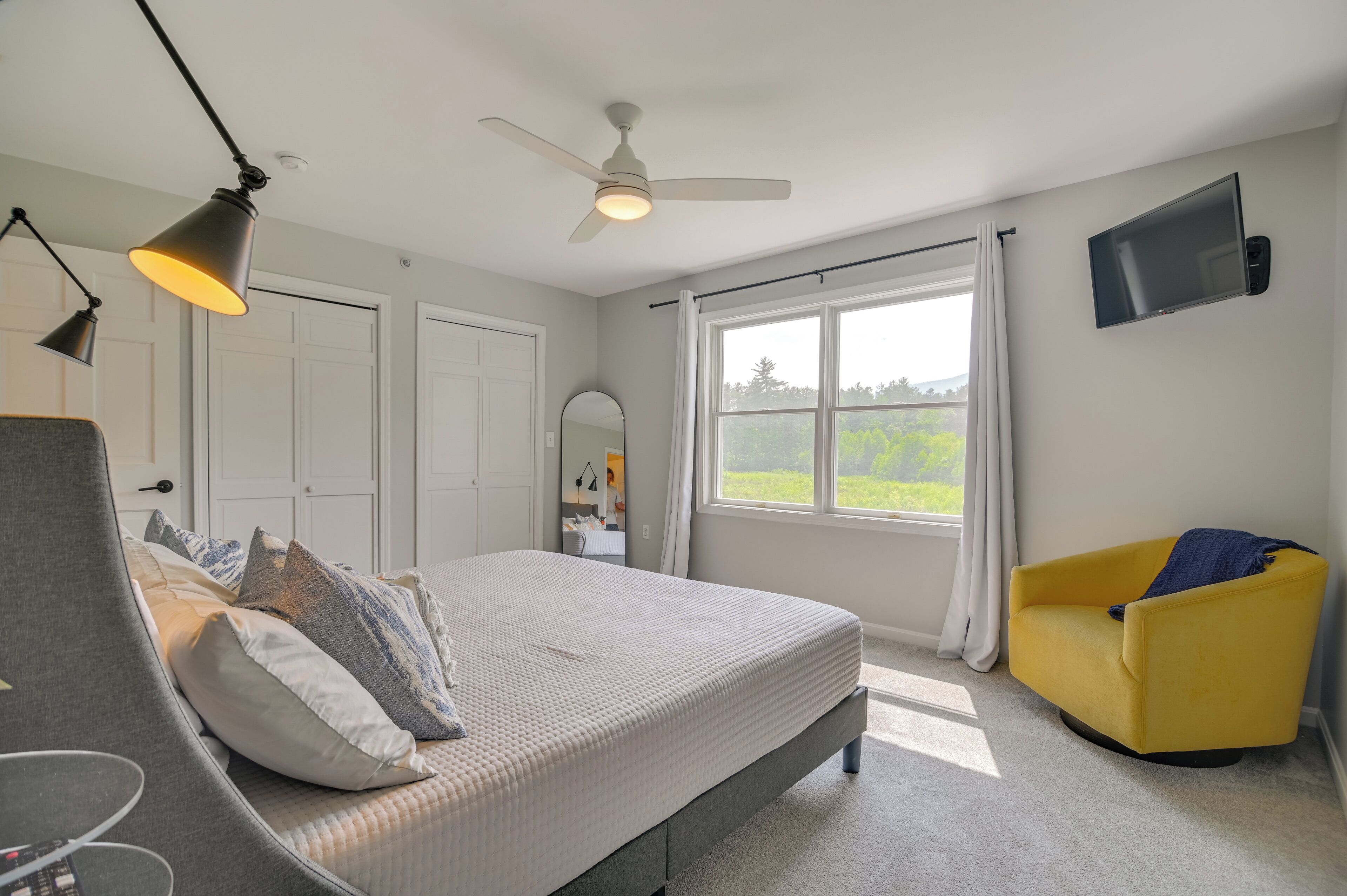 A bedroom with a bed, a chair, a ceiling fan, a TV, and a window with a view of greenery.